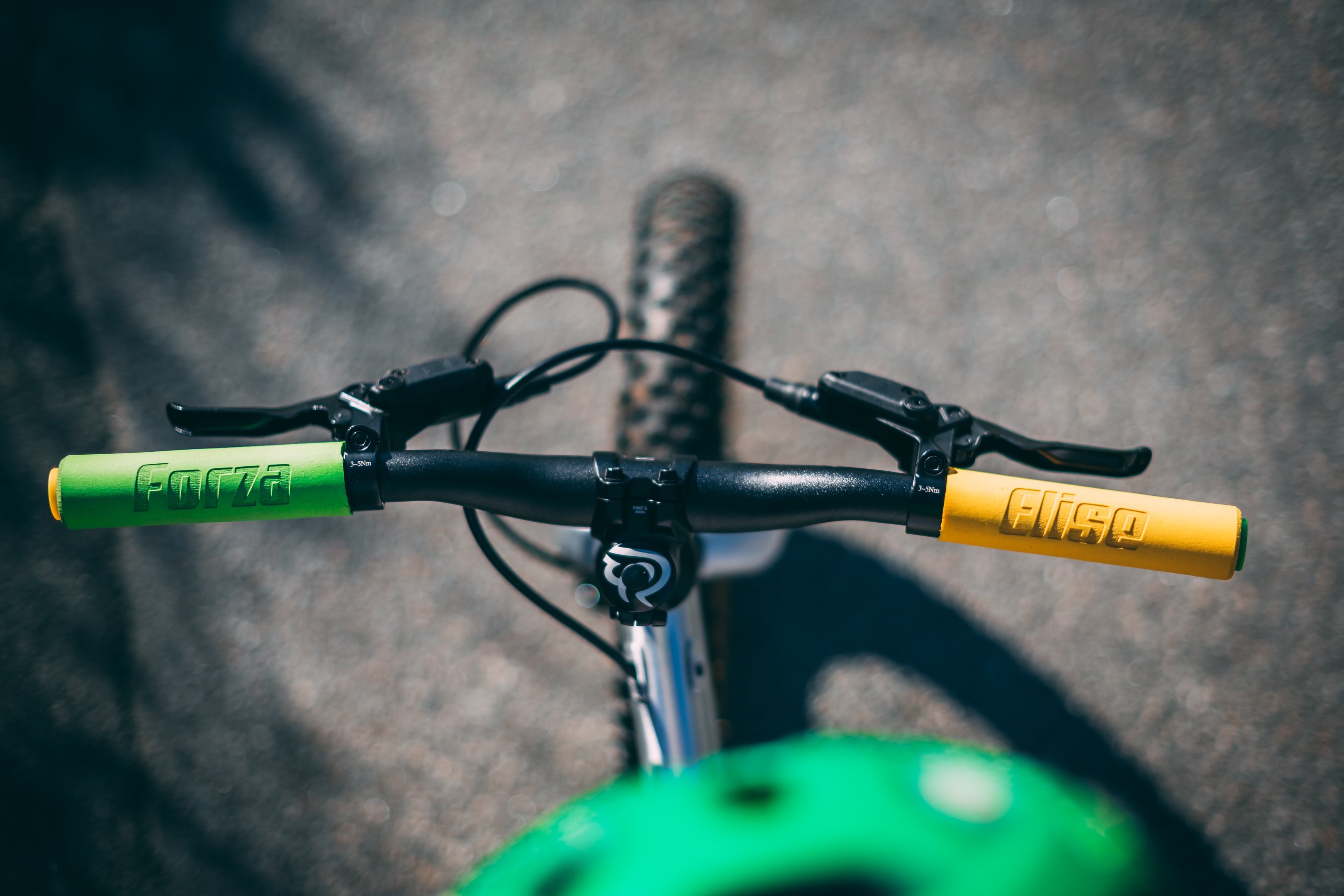 Aerial photo looking down on small bicycle handlebar. Two bright colored ESI grips stand out to an asphalt background. The left grip is green and is engraved with the word "Forza". The right grip is yellow and reads "Elise". 