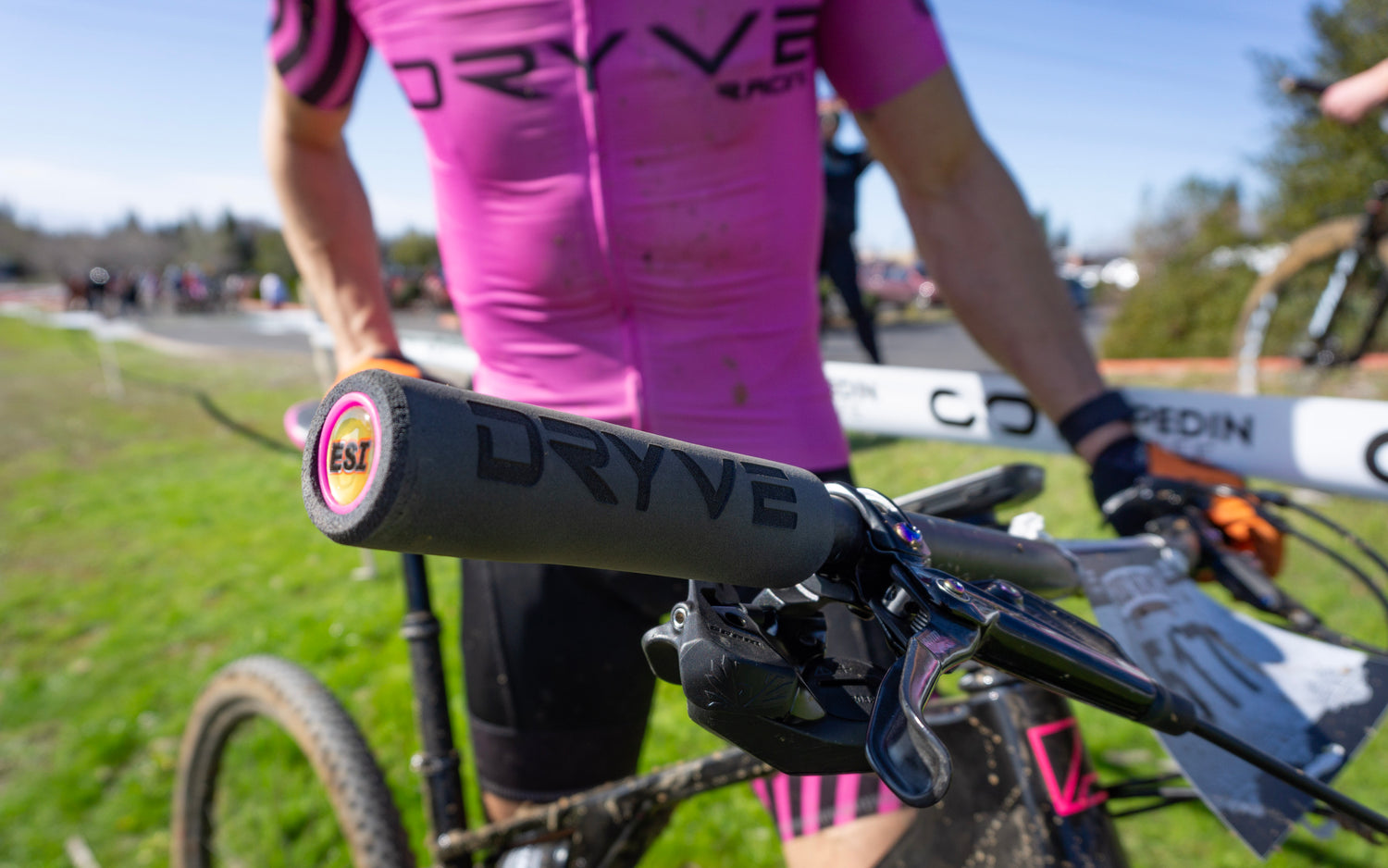 An engraved black esi grip is the focal point of the image. The engraved grip reads "Dryve" in the brands logo. Out of focus in the background is an athlete at a race dressed in a bright pink cycling kit from the team Dryve.